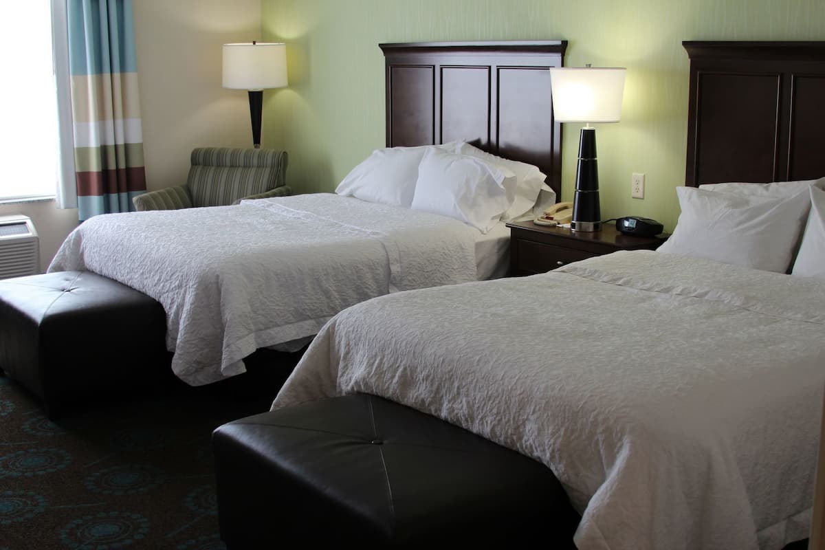 A hotel room with two beds, a chair, a nightstand with a lamp, and a window with curtains.