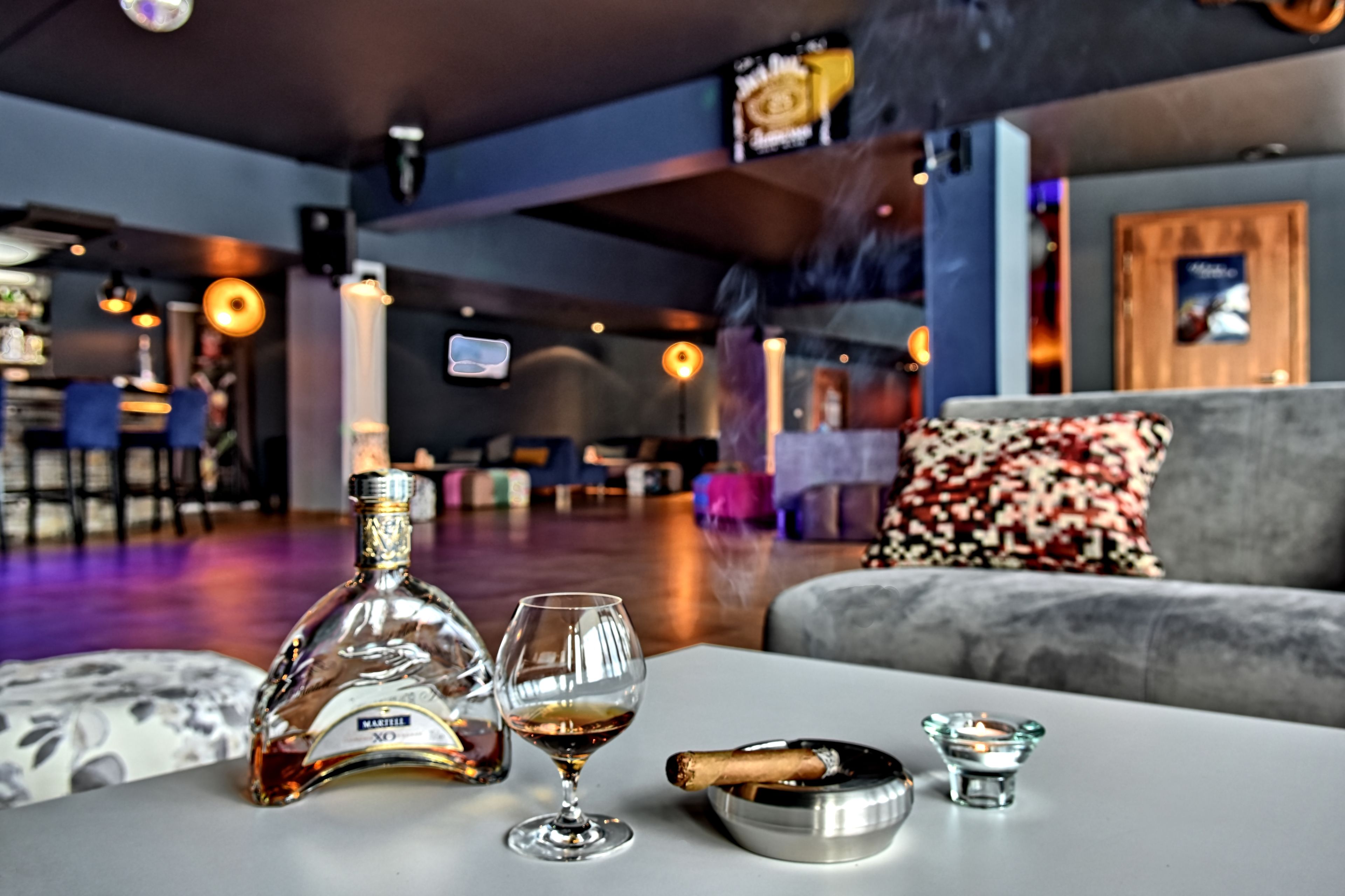 A bar area with a bottle of liquor, a glass, a cigar, and a ashtray on a table.