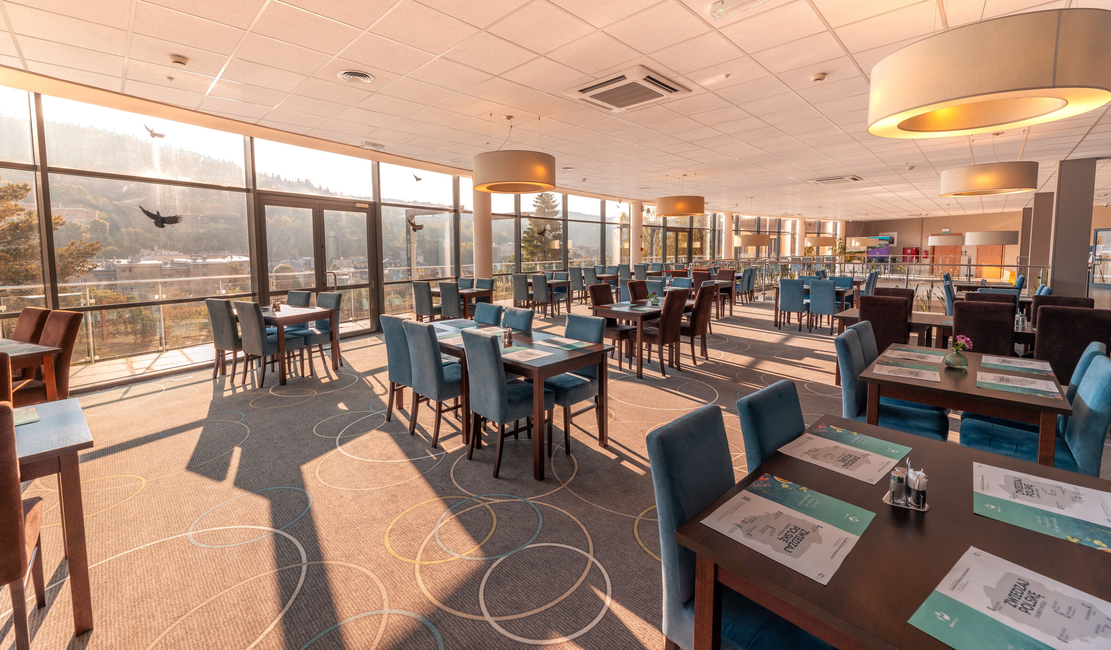 Ein geräumiges Restaurant mit hohen Decken, großen Fenstern, die einen Blick auf die Stadt bieten, und moderner Einrichtung.