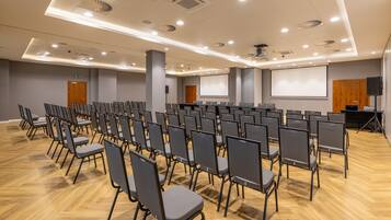 Un salón de conferencias con filas de sillas, una pantalla de proyector y un espacio para presentaciones.