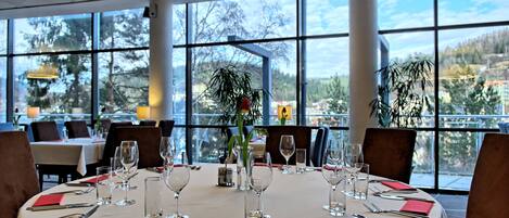 A dining area with round tables set for a meal, large windows offering a view of trees and buildings, and a modern interior design.