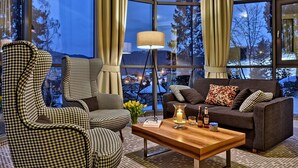 A cozy living room with a houndstooth armchair, a dark sofa with patterned cushions, a wooden coffee table with a candle, and a floor lamp. - Hotel Krynica Conference & SPA (Krynica-Zdroj)