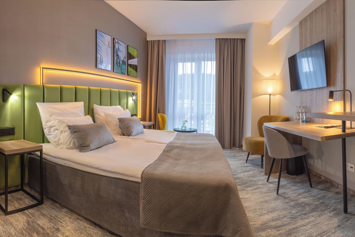 A hotel room with a bed, desk, chair, TV, and artwork on the wall.