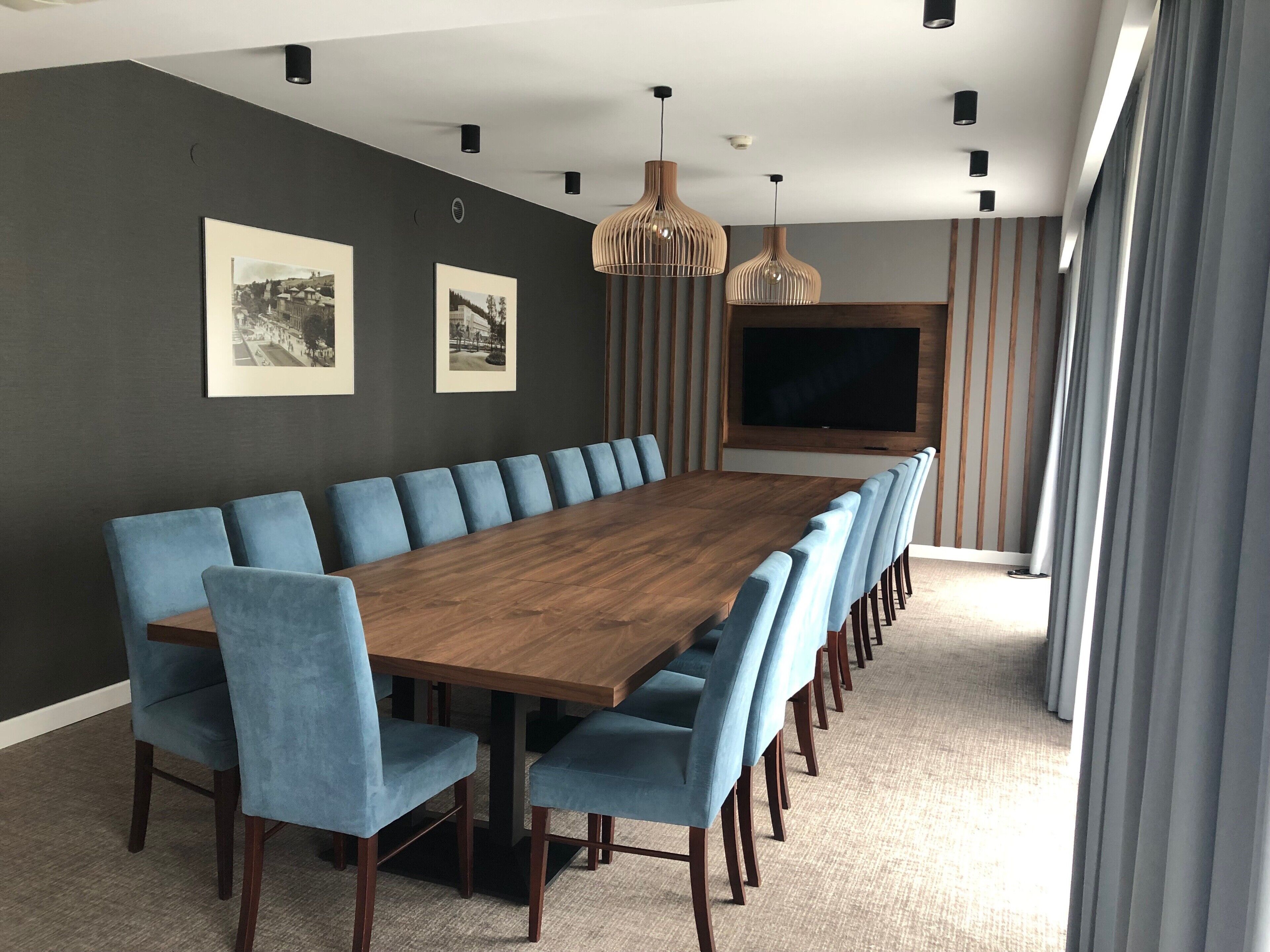 A conference room with a long wooden table, blue upholstered chairs, pendant lighting, and a large flat-screen TV mounted on the wall.