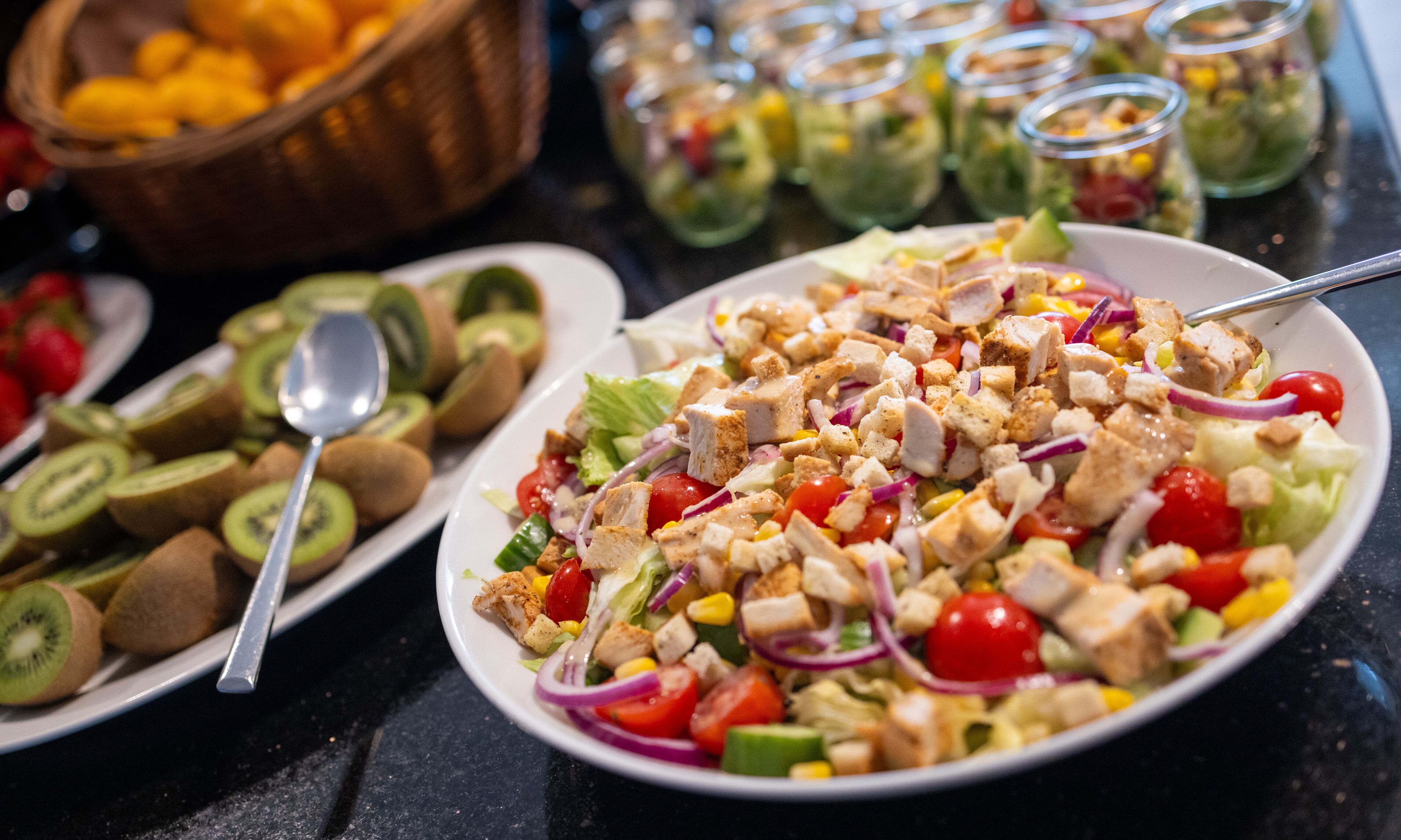 Une table de buffet proposant divers plats, notamment une salade composée de poulet, de légumes et accompagnée de tranches de kiwi.