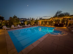 A swimming pool with lounge chairs and string lights, surrounded by palm trees and a building in the background.