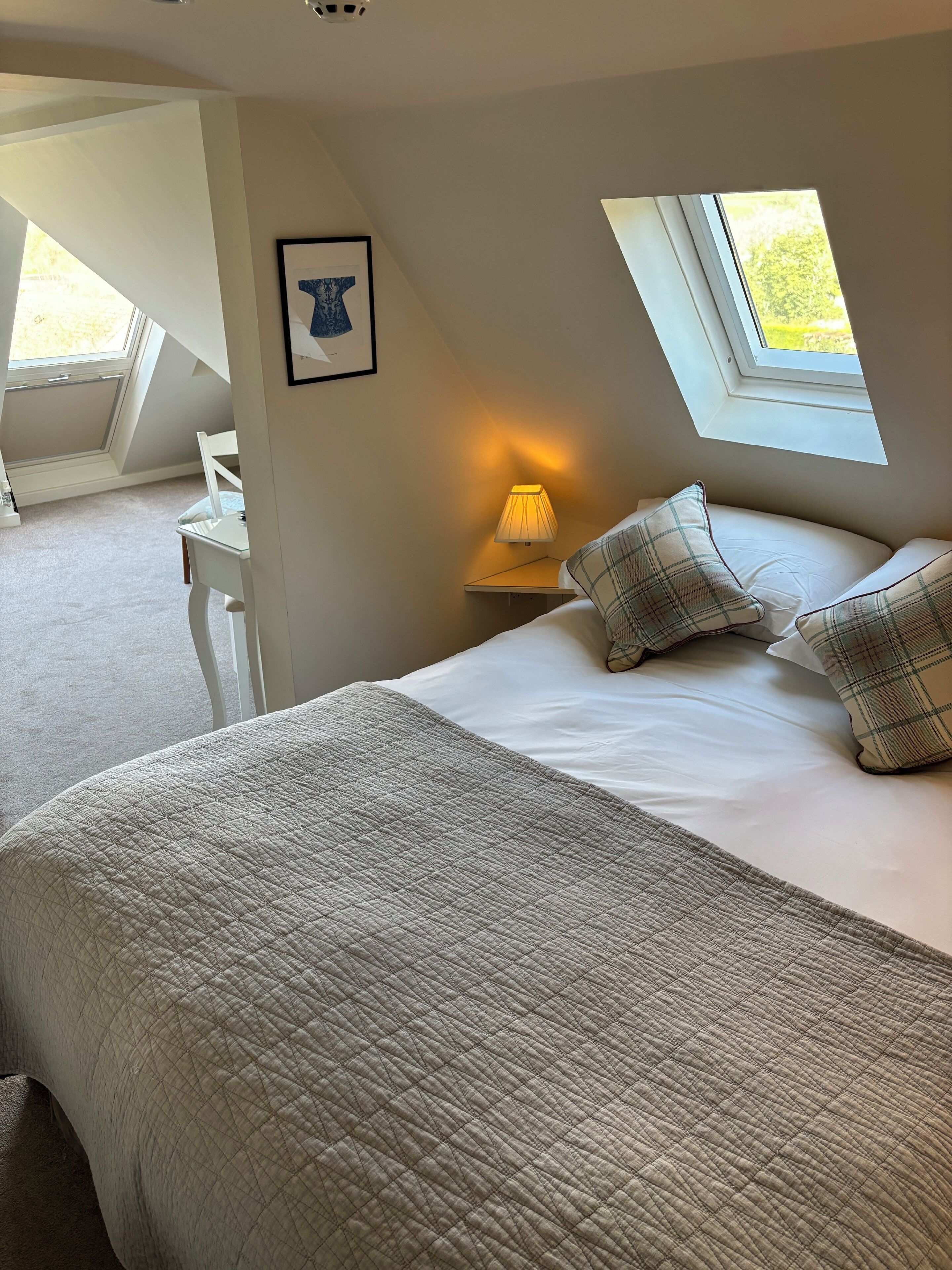 Une chambre avec un grand lit, un bureau et une chaise, une lucarne et un tableau encadré au mur.
