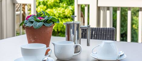 A table with a coffee pot, two cups, and a saucer, a potted plant, and a wicker chair.