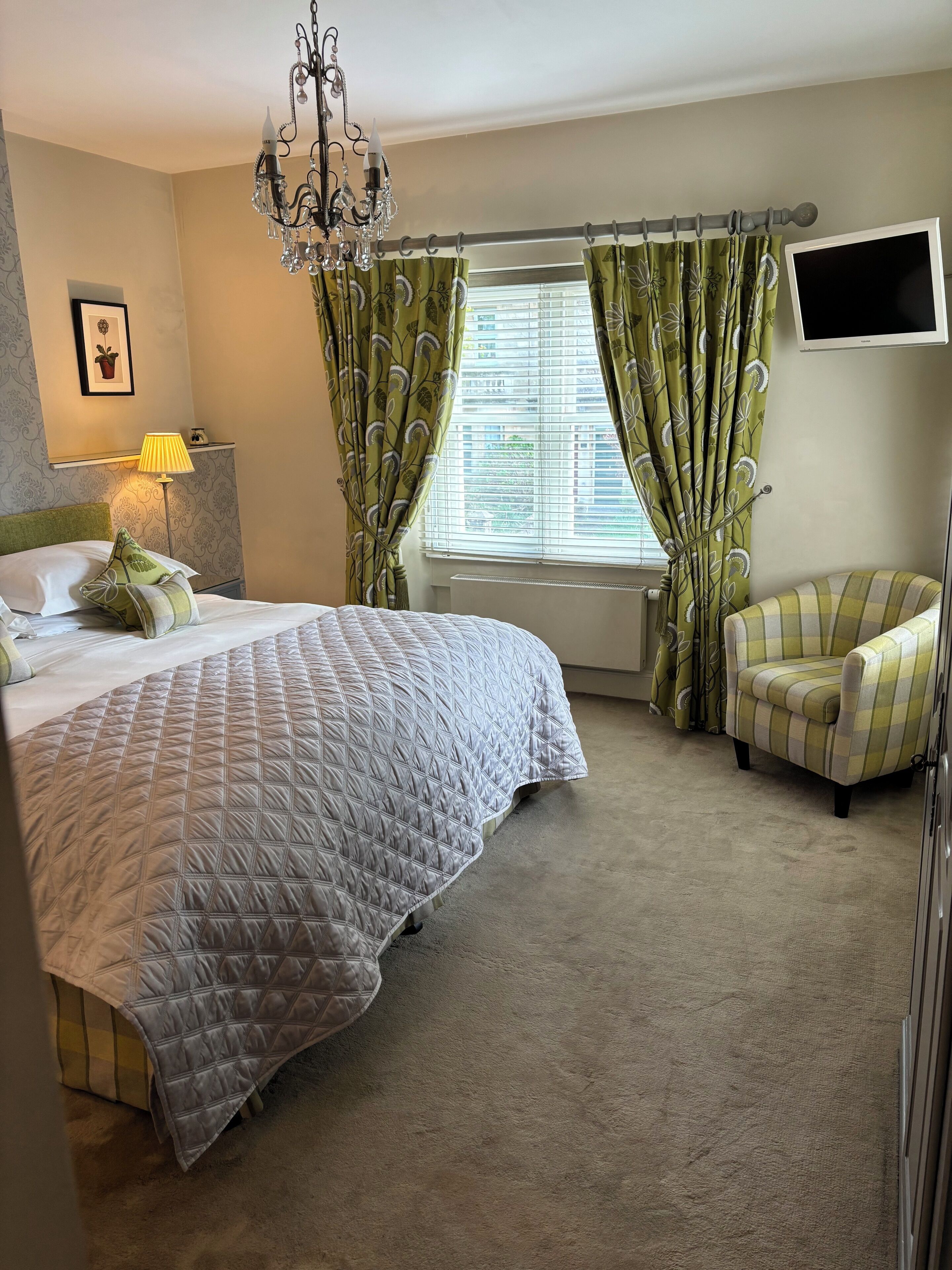 A bedroom with a large bed, a chandelier, a chair, and a TV mounted on the wall.