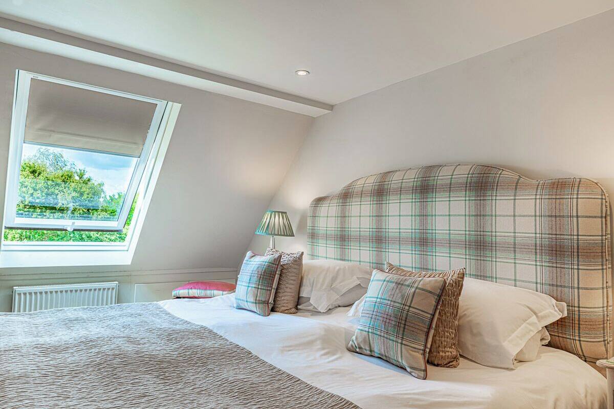 A bedroom with a large bed, a window with a view of greenery, a bedside table with a lamp, and a plaid-patterned headboard.