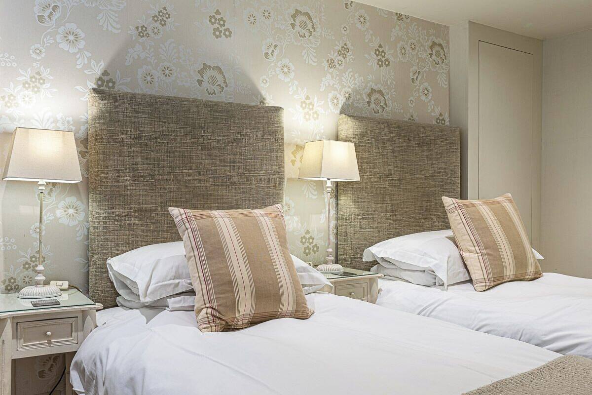 A hotel room with two beds, each with a headboard, white bedding, and striped pillows. There are bedside tables with lamps and a small decorative item on one table.