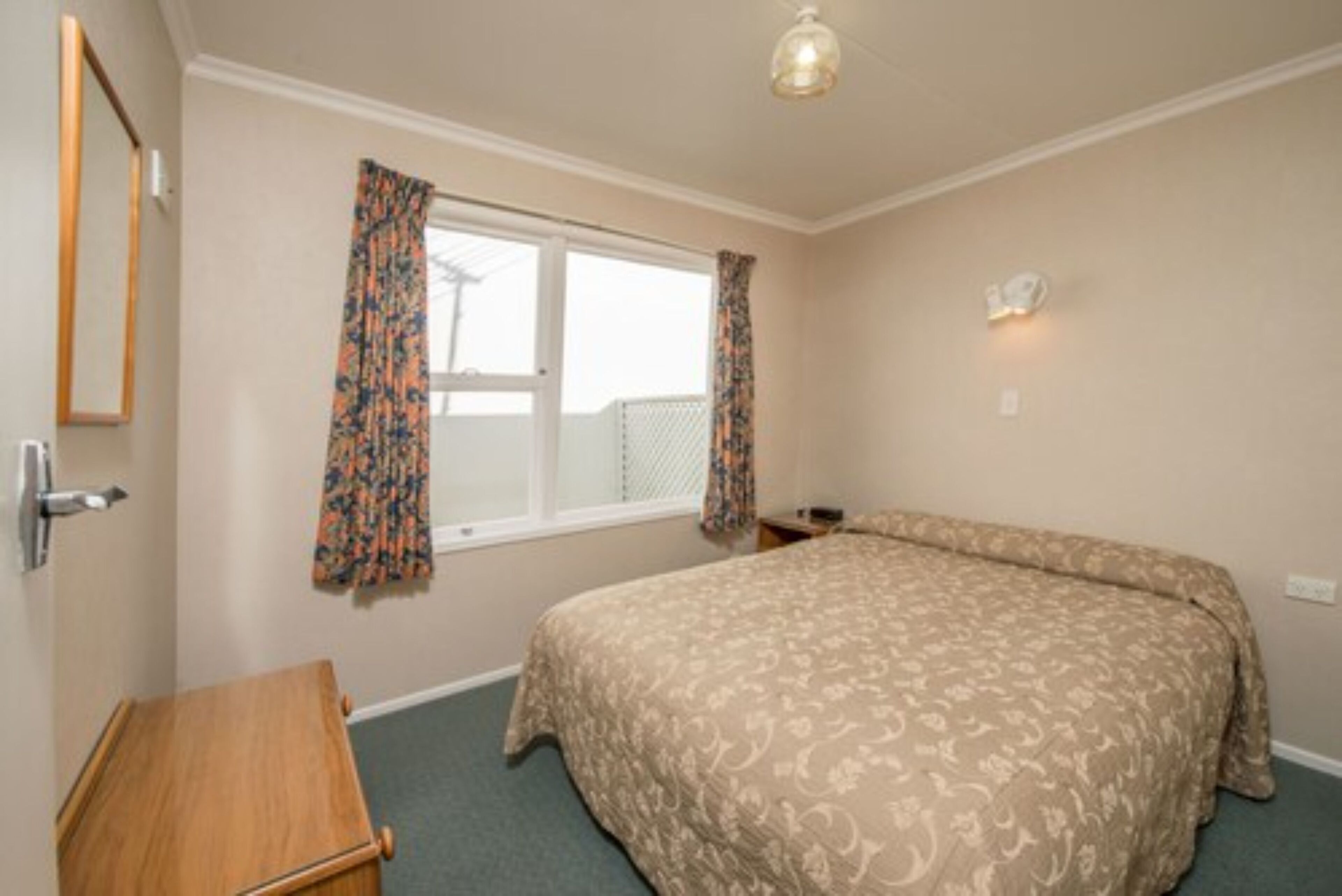 A bedroom with a bed, a wooden dresser, a window with curtains, and a wall-mounted light.