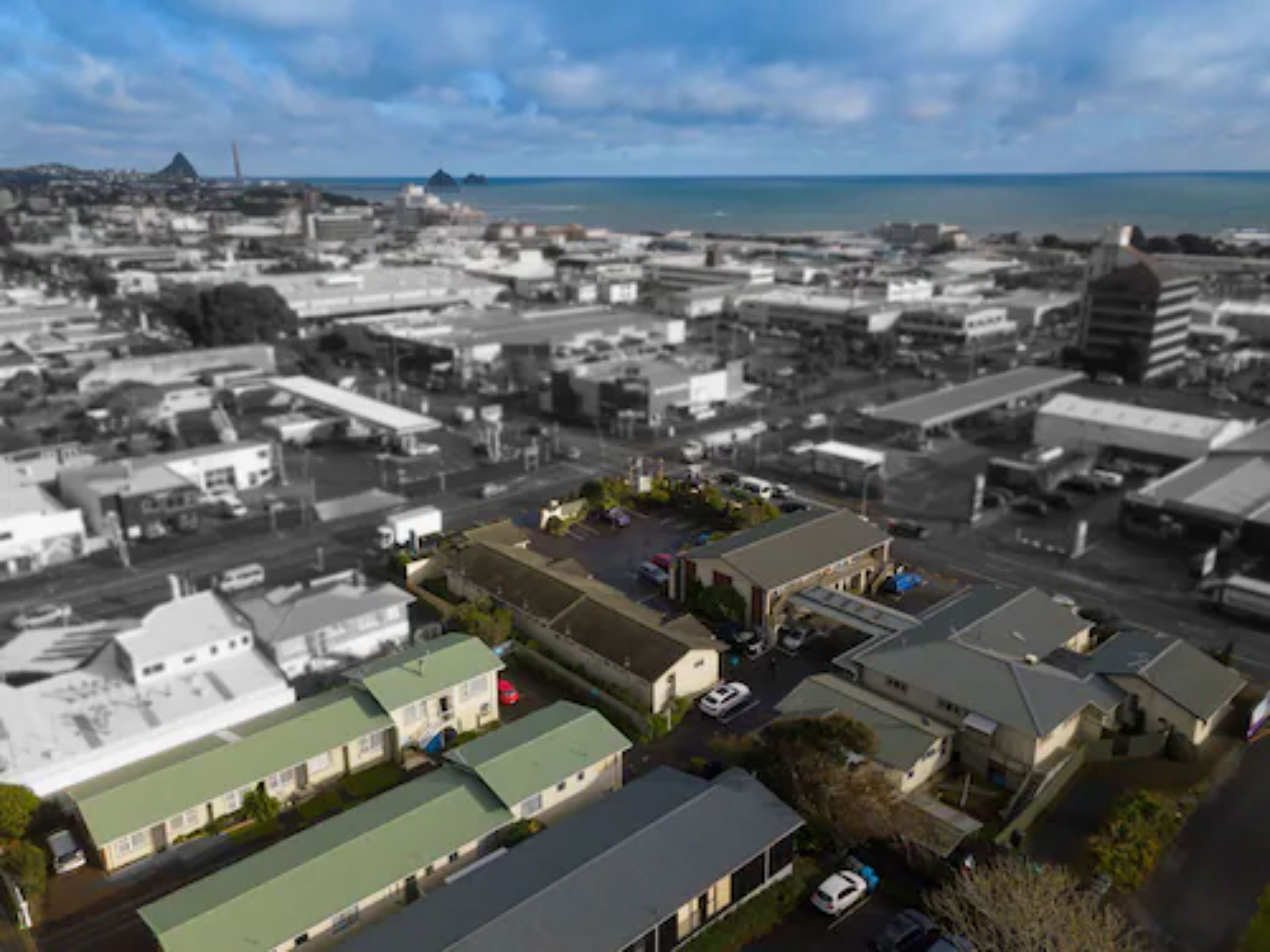 A black and white aerial view of a coastal town with a mix of residential and commercial buildings, a prominent mountain in the background, and a clear view of the sea.