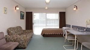 Et lille, godt oplyst rum med seng, sofa, bord og to stole. Der er et vindue med gardiner, en lampe på væggen og et billede.
