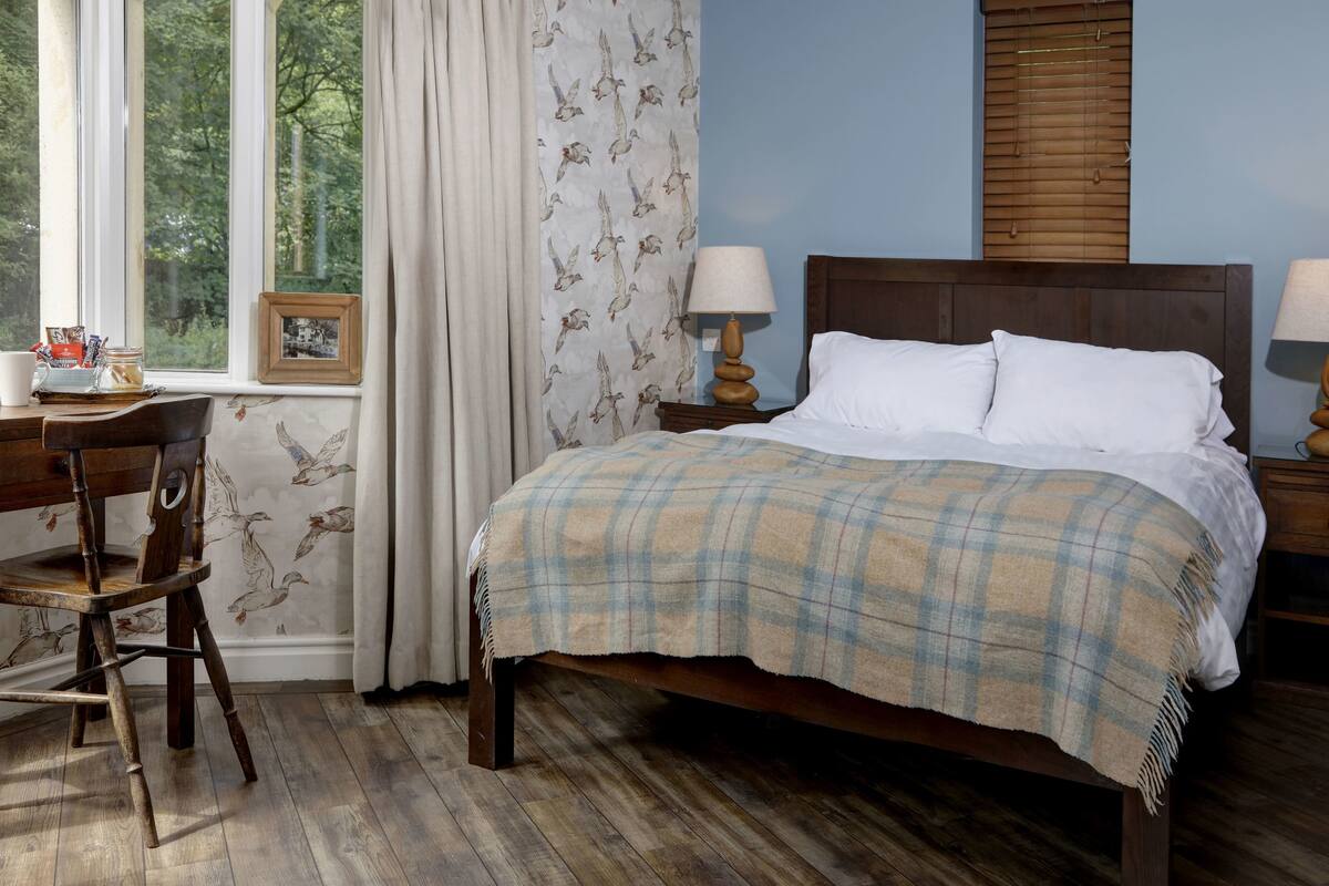 Quarto com cama de madeira, duas mesas de cabeceira com luminárias, escrivaninha de madeira com cadeira, janela com cortinas e parede com padrão de pássaros.