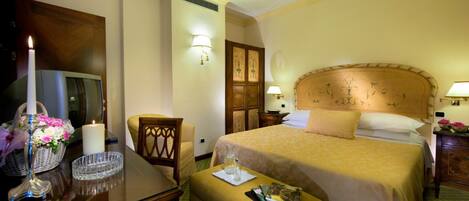 Habitación de hotel con una cama grande, cabecera de madera, escritorio con lámpara y una mesita con vela y flores.