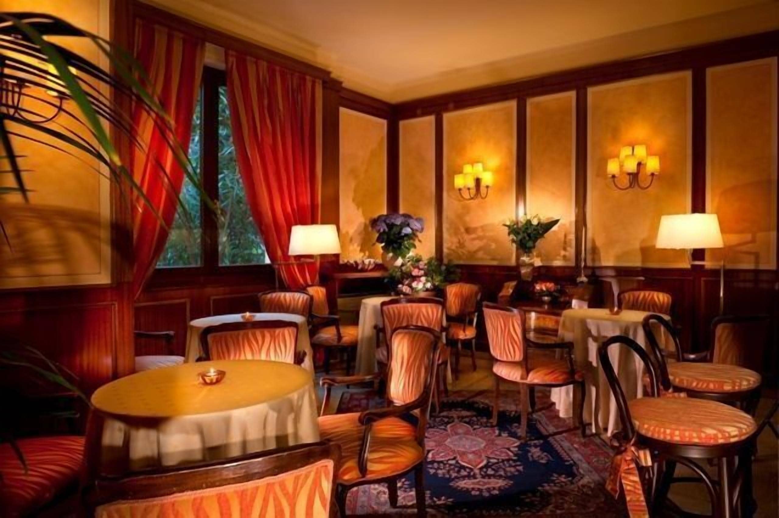 A hotel room with a large window, red curtains, a round table with a candle, several chairs, and wall-mounted lamps.