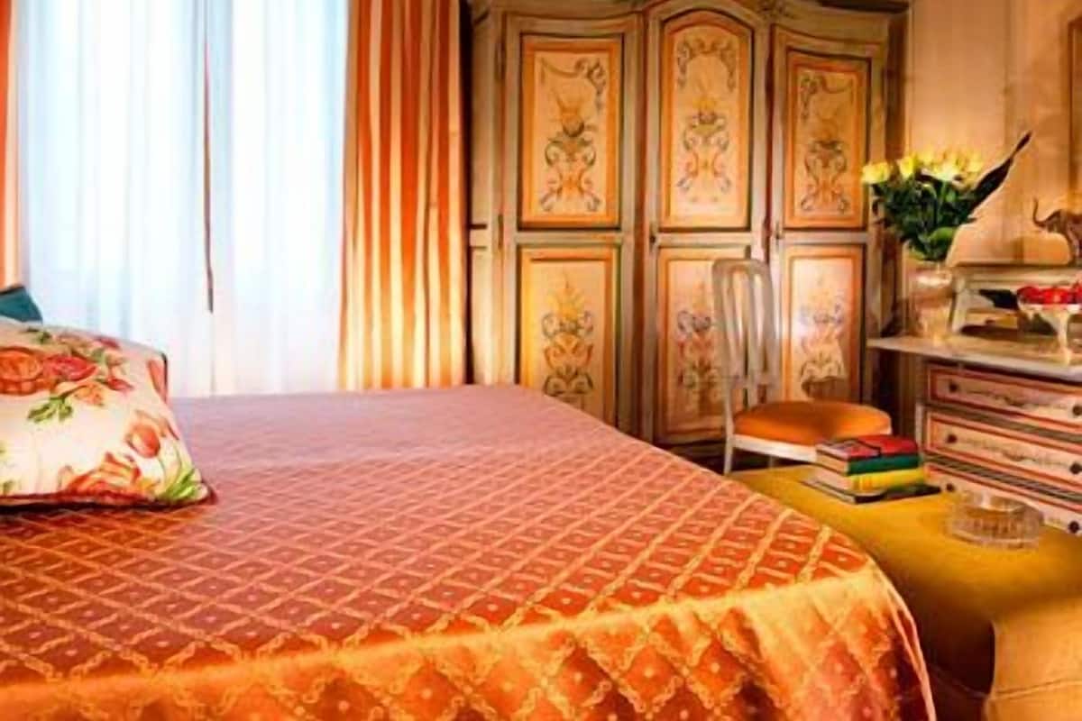 A bedroom with a large bed, a wooden wardrobe, and a small table with books and a vase of flowers.