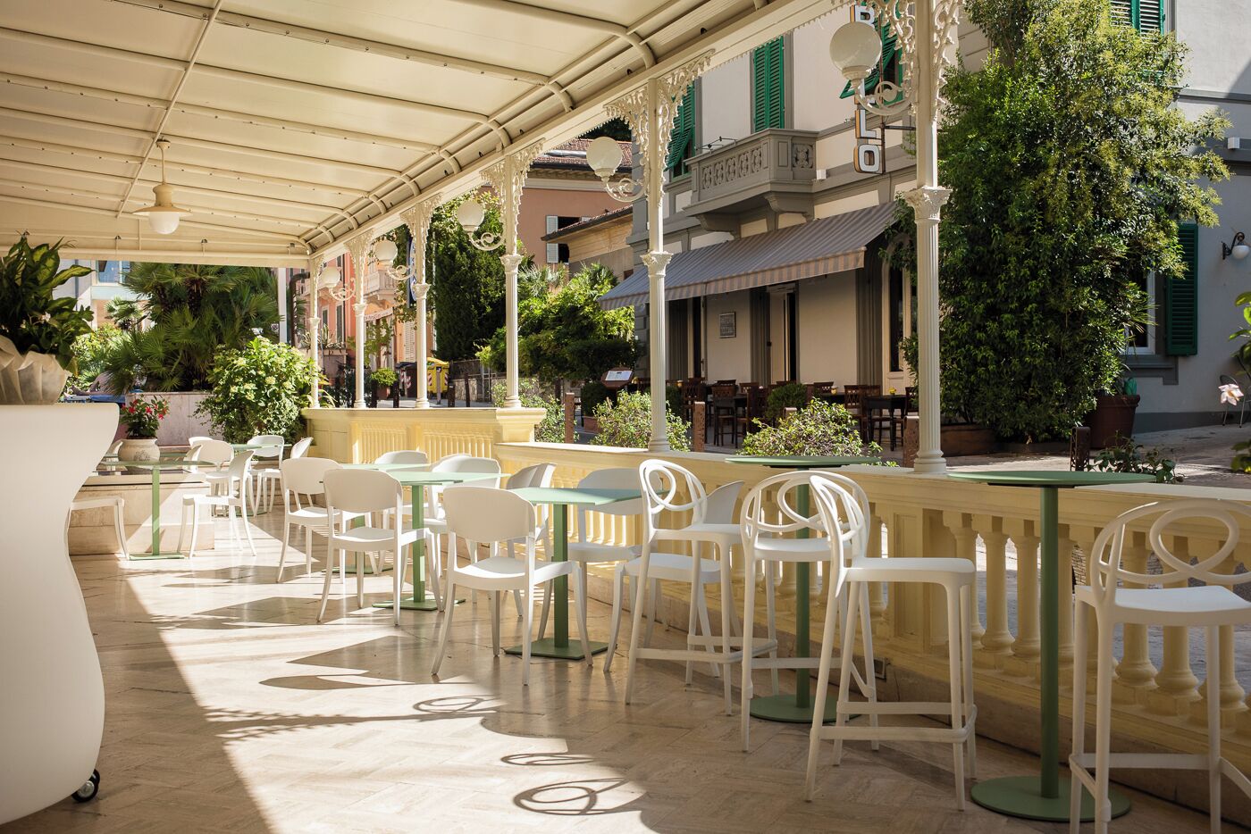 Un café en plein air avec des chaises et des tables blanches, abrité par un auvent et entouré de verdure et d’immeubles.