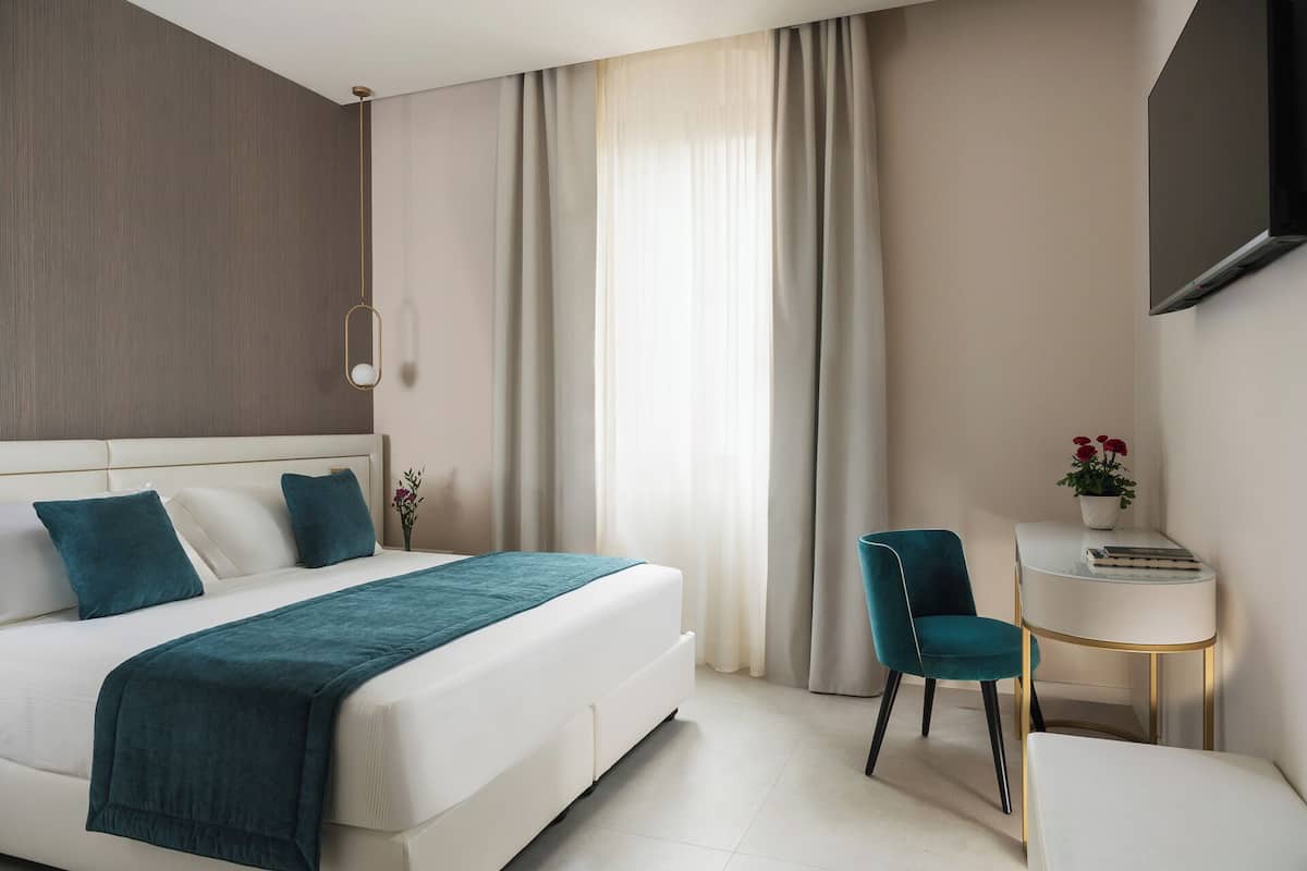 A modern hotel room with a large bed, a bedside table with a lamp, a chair, and a small table with a plant.