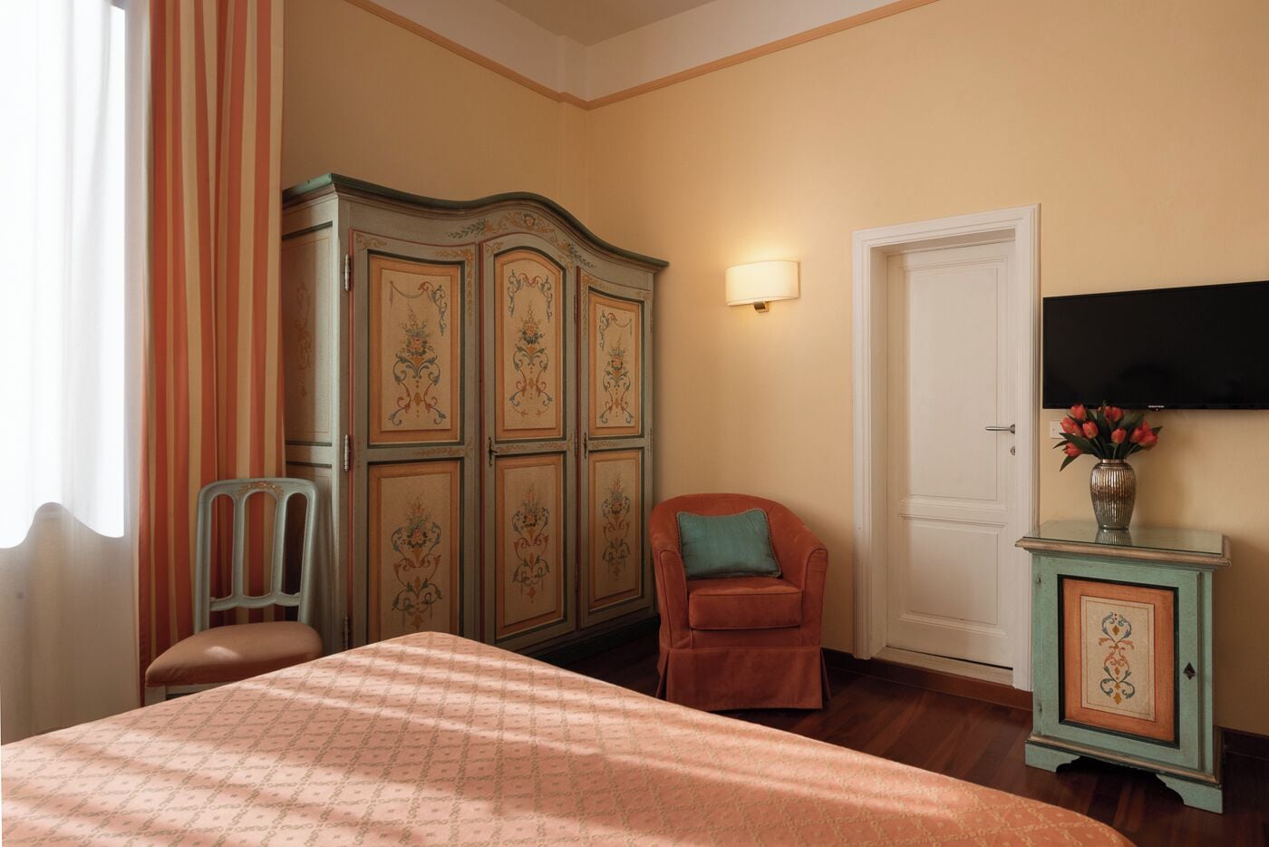 A hotel room with a bed, a chair, a wardrobe, a television, and a small cabinet with a vase of flowers.