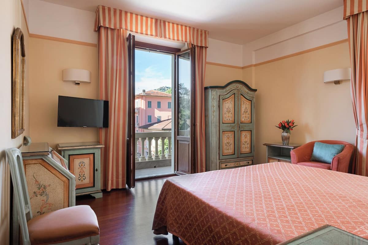 A hotel room with a bed, a television, a wardrobe, a chair, and a balcony with a view of buildings.