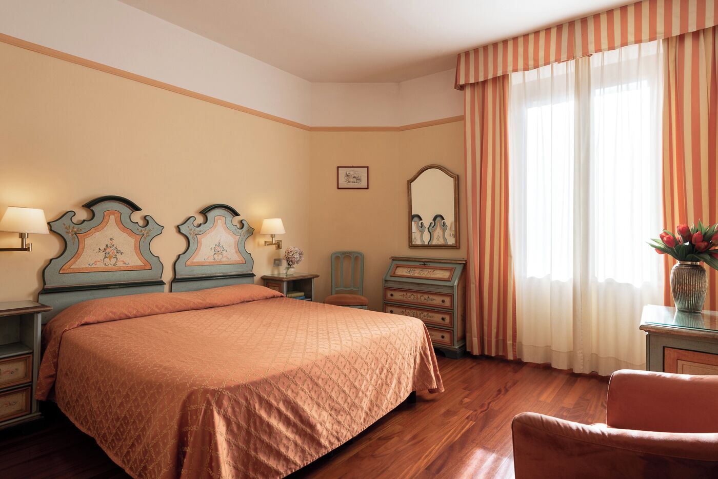 A hotel room with a large bed, two bedside tables with lamps, a dresser, a mirror, and a window with striped curtains.