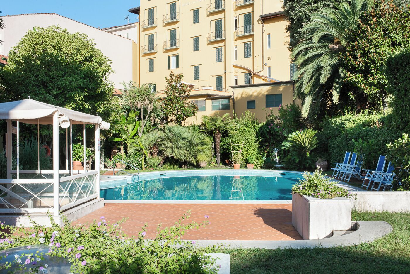 A swimming pool surrounded by greenery, a covered seating area, and a multi-story building with balconies.