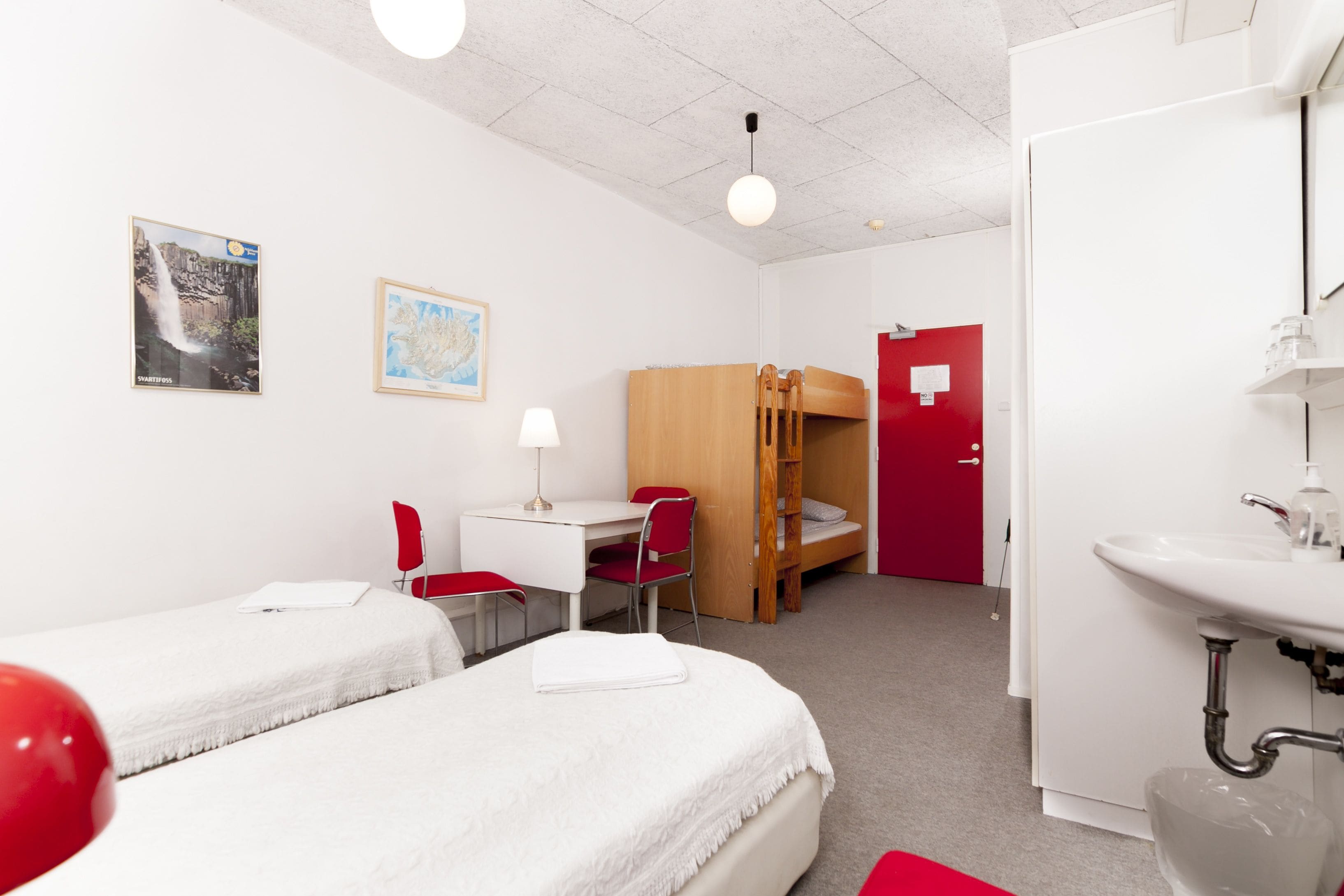 A hotel room with two beds, a desk, a chair, a bunk bed, a sink, and a red door.
