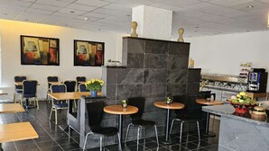 Un restaurante moderno con suelos de baldosas negras, mesas de madera y sillas negras. Hay dos cuadros en la pared, una chimenea de mármol negro y una barra con equipo de cocina.
