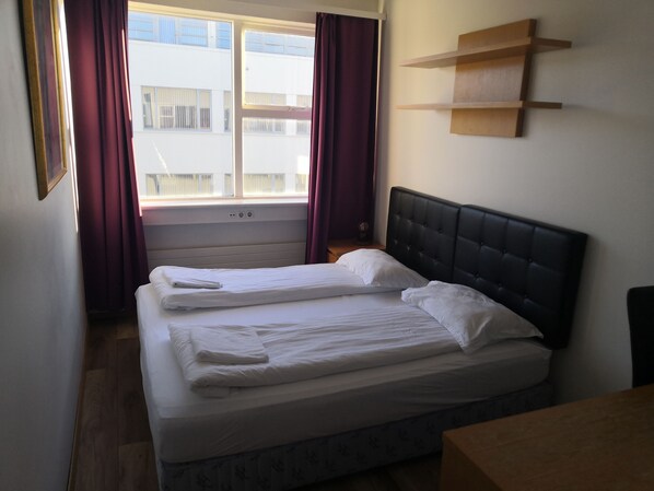 Un lit double avec une tête de lit, une fenêtre avec des rideaux et une étagère en bois fixée au mur.