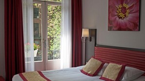 A hotel room with a bed, red curtains, a window with a view of greenery, and a wall-mounted lamp.