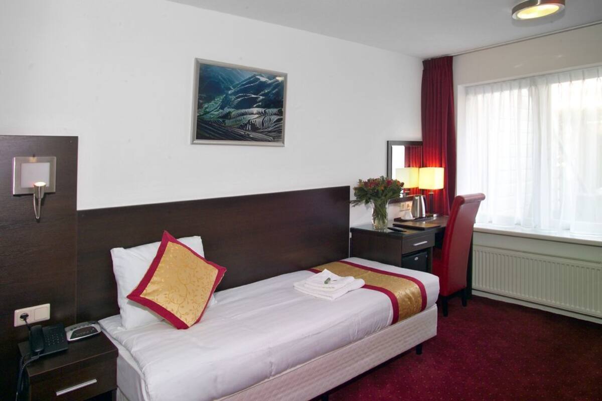 A hotel room with a bed, a desk, a chair, a telephone, a lamp, a painting, and a window with curtains.
