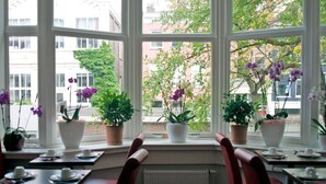 A dining area with tables and chairs, large windows with potted plants, and a view of buildings and trees outside.
