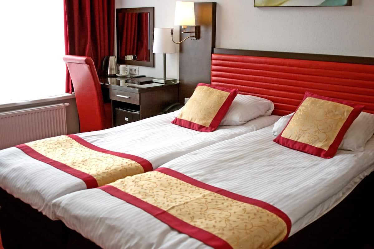 A hotel room with two beds, each with red and gold bedding, a red headboard, a desk with a chair, a lamp, and a mirror.
