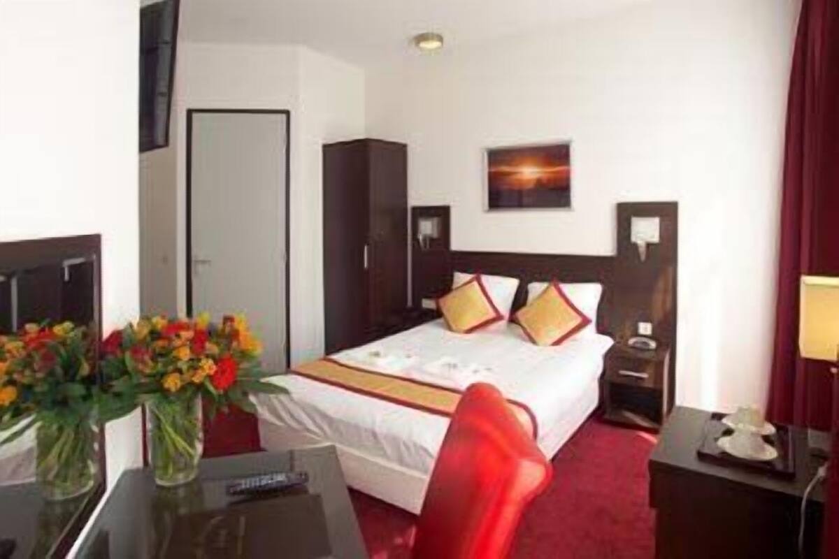 A hotel room with a bed, a desk, a chair, a nightstand, a lamp, and a vase with flowers.