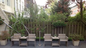 A row of wooden benches with potted plants on a paved patio area, surrounded by a wooden fence and greenery.
