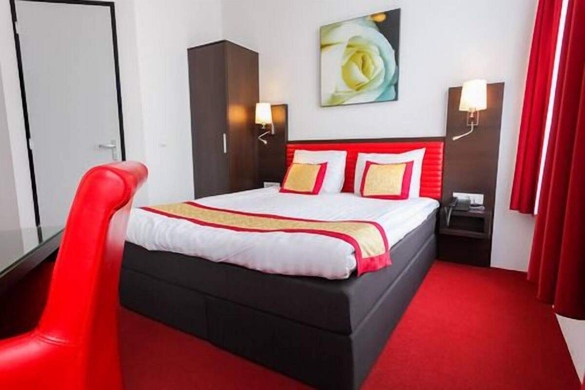 A hotel room with a large bed, red and white bedding, a red chair, a painting of a rose, and two wall-mounted lamps.