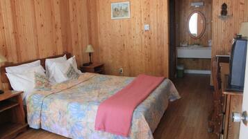 A wooden-paneled room with a bed, a nightstand, a lamp, a painting, a mirror, and a television.