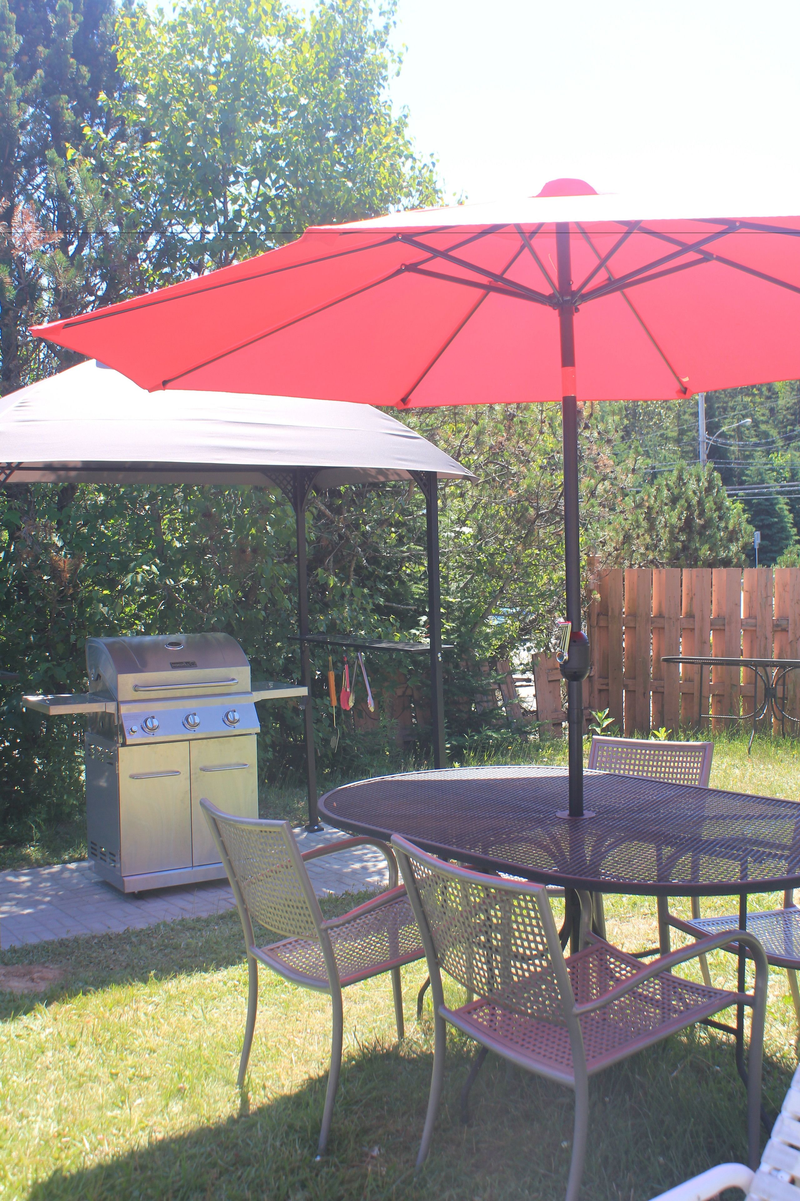 An outdoor patio with a metal table and chairs, a red umbrella, and a barbecue grill.