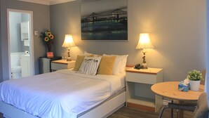 A hotel room with a large bed, two bedside tables with lamps, a small round table with a chair, a painting of a bridge, and a bathroom visible through an open door. - Motel des Pays d'en Haut (Val David)