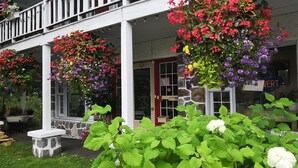 A building with a white exterior, a stone foundation, and a porch with hanging flower baskets. - Motel des Pays d'en Haut (Val David)