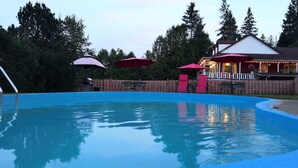 A swimming pool with a wooden deck, surrounded by a wooden fence and trees. There are umbrellas and outdoor seating. - Motel des Pays d'en Haut (Val David)