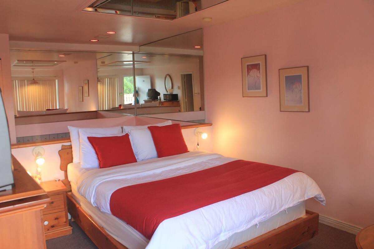A neatly made bed with a red and white bedspread, a large mirror above the bed, and two framed pictures on the wall.