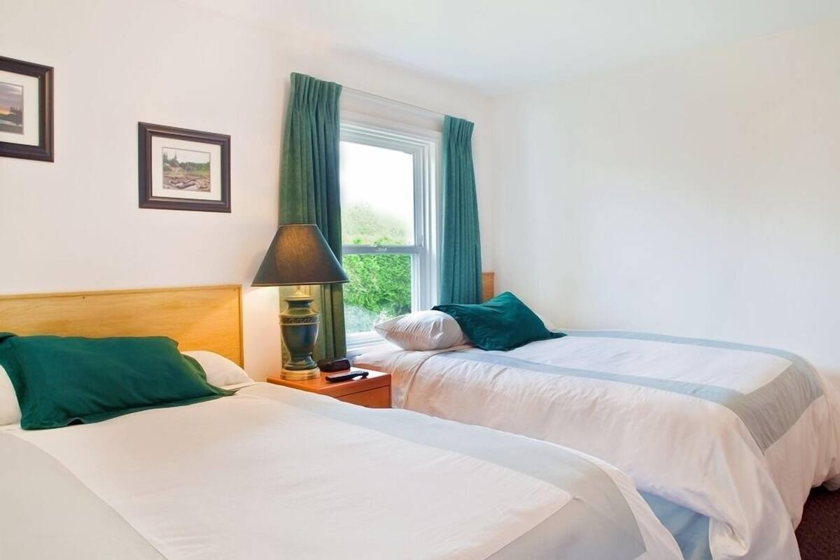 A hotel room with two beds, a nightstand with a lamp, and a window with curtains.