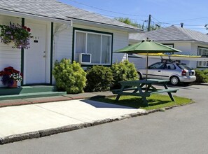 A single-story motel with a green picnic table and umbrella, a white car parked in front, and a small garden area with potted plants. - Somass Motel (Port Alberni)