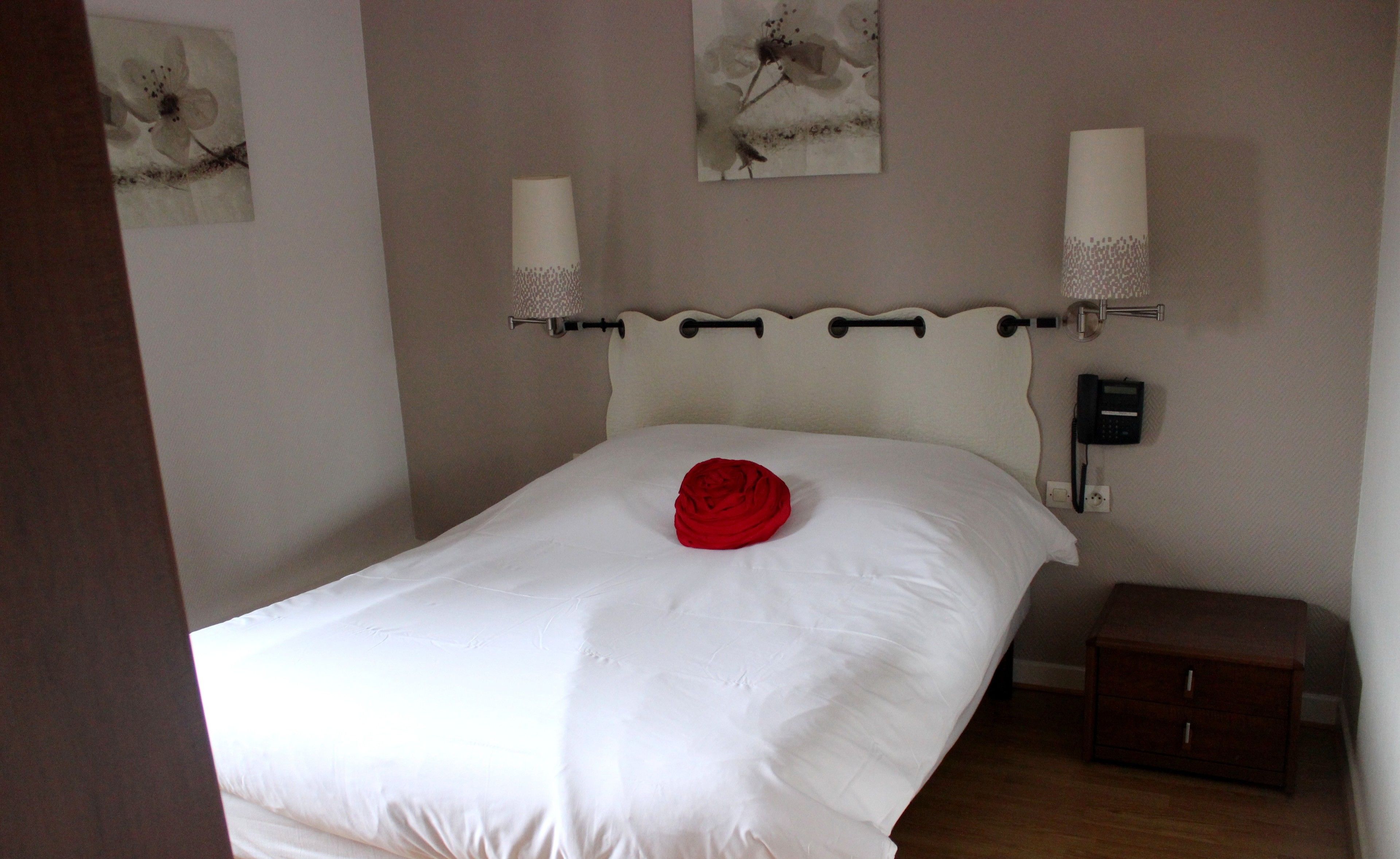 A neatly made bed with a red pillow, a bedside table, a wall-mounted phone, and two wall sconces.