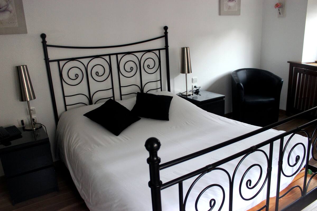 A neatly made bed with black headboard and footboard, a bedside table with a lamp, a chair, and a small table with a lamp.
