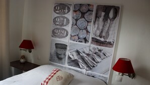 Una cama bien hecha con un cojín a cuadros, dos mesitas de noche con lámparas y una obra de arte en la pared que representa utensilios de cocina y números.