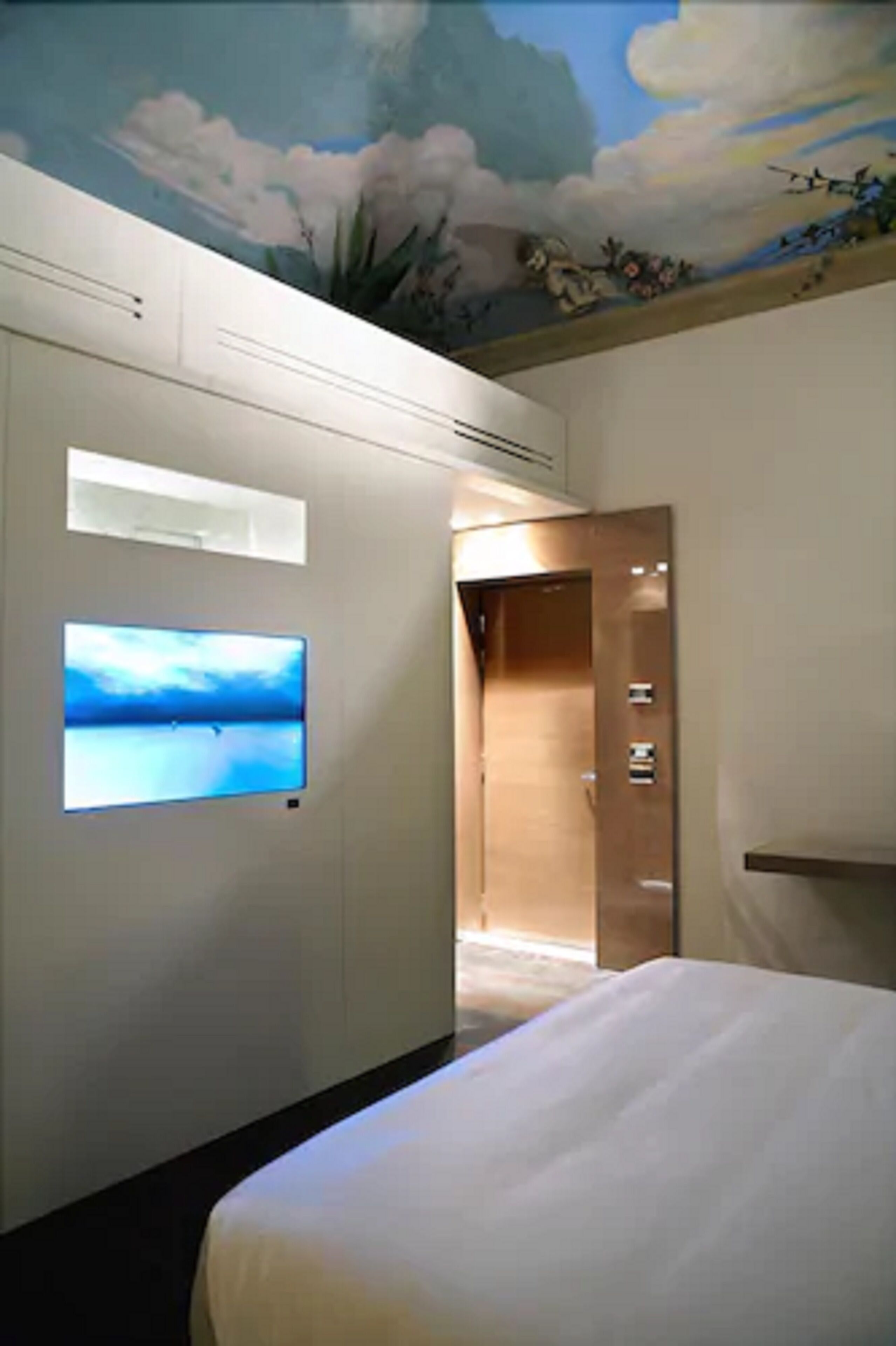 Een hotelkamer met een bed, een tv aan de muur, een schilderij op het plafond en een deur naar een andere kamer.